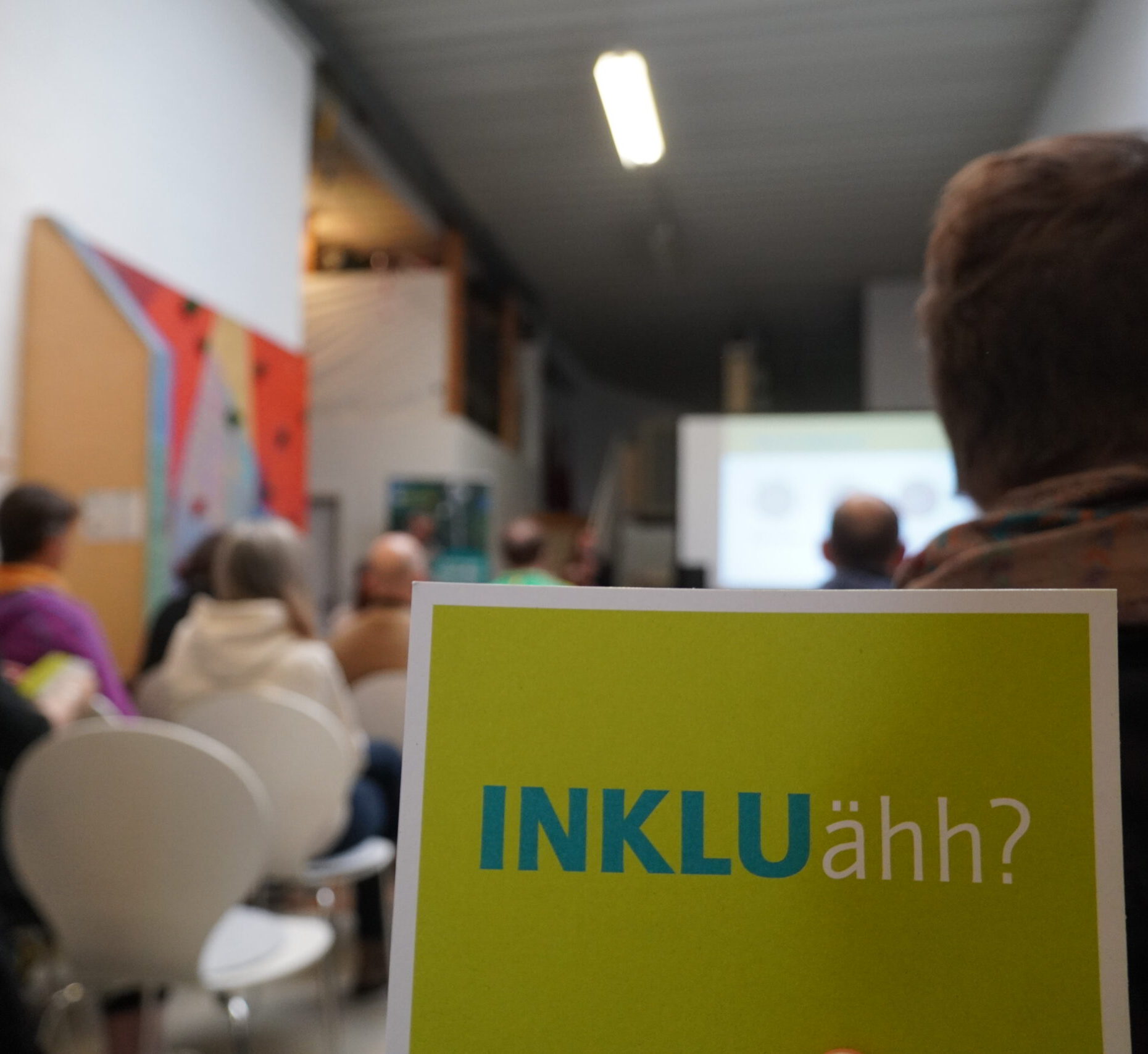 Im Vordergrund ist eine Postkarte auf der "INKLUähh?" steht. Im Hintergrund sitzen Menschen vor einer Leinwand. Ein Stuhl ist frei.