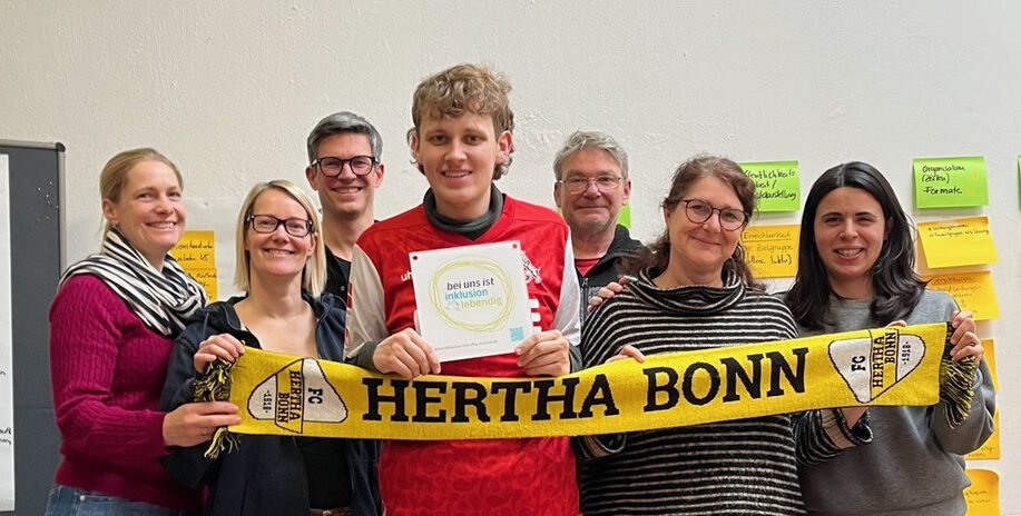 Sieben Menschen steen hinter einem ausgebreiteten Schal von ihrem Verein Hertha Bonn und ein Mensch in der Mitte hält die Plakette von dem Label "Hier ist Inklusion lebendig" hoch.